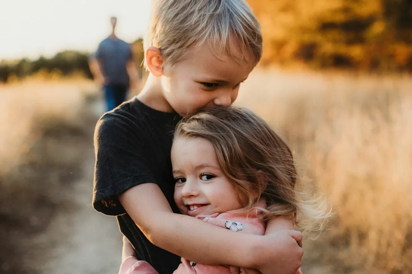 Teaching kindness and respect to children at home Ein Junge umarmt ein Mädchen lächelnd auf einem Feld. Im Hintergrund ist eine Person zu sehen.